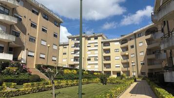 Un immeuble résidentiel à plusieurs étages, doté de balcons, d’un chemin central et d’un jardin bien entretenu avec différents arbustes et fleurs.
