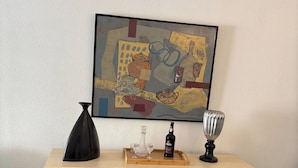 Une pièce comprenant un meuble en bois, un tableau abstrait encadré, un vase noir, une bouteille de spiritueux et un verre posé sur le meuble.