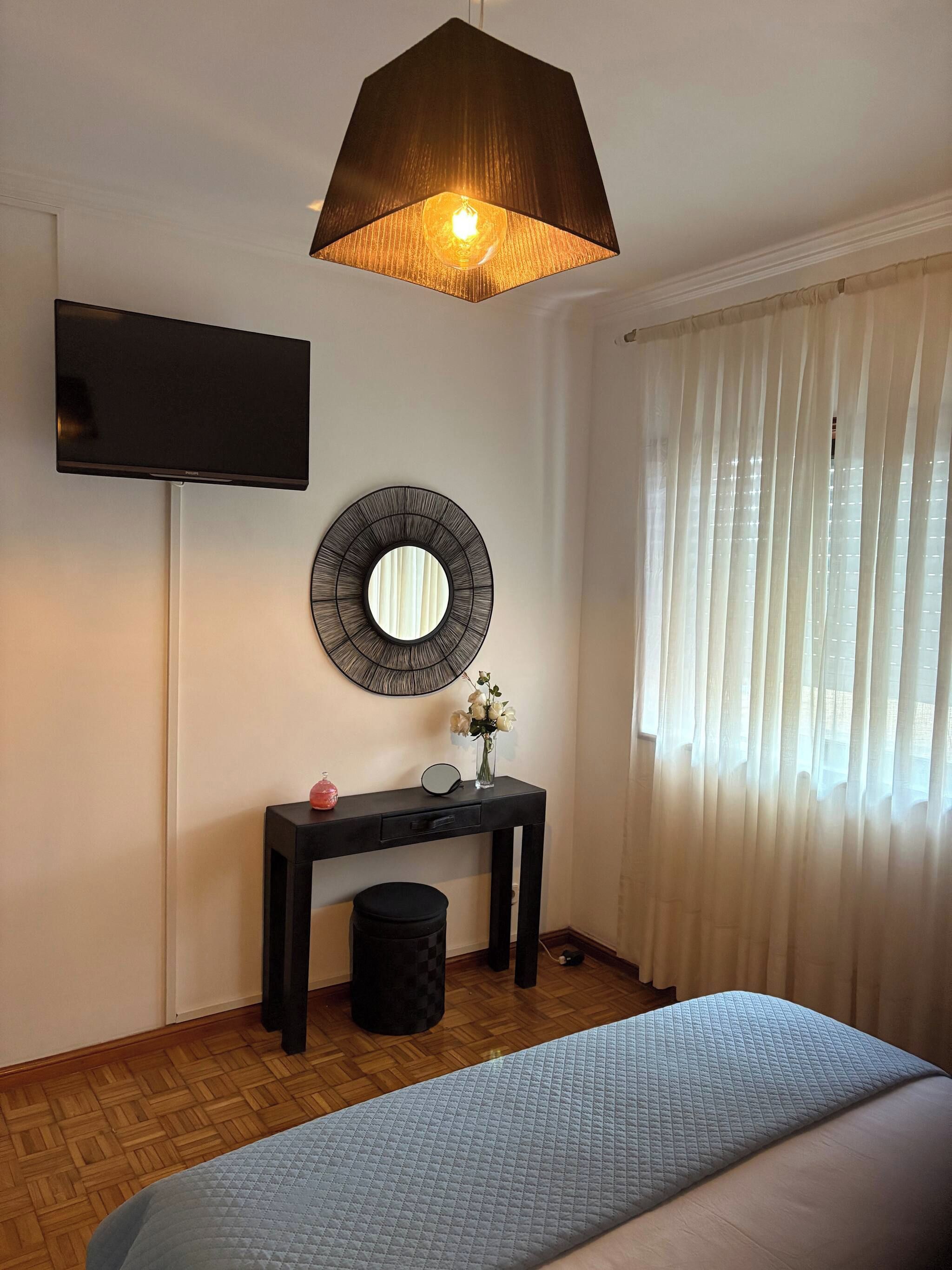 A bedroom with a bed, a bedside table with a mirror, a small vase with flowers, a television mounted on the wall, and a window with curtains.