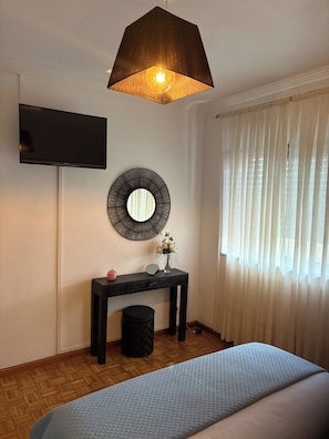A bedroom with a bed, a bedside table with a mirror, a small vase with flowers, a television mounted on the wall, and a window with curtains.