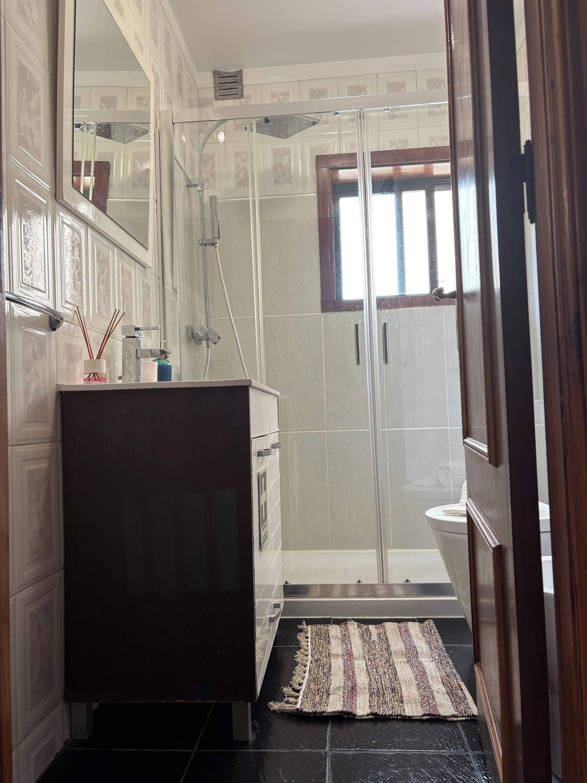 Une salle de bain dotée d’une douche à parois de verre, d’un meuble-lavabo en bois foncé et d’un tapis à motifs sur le sol.