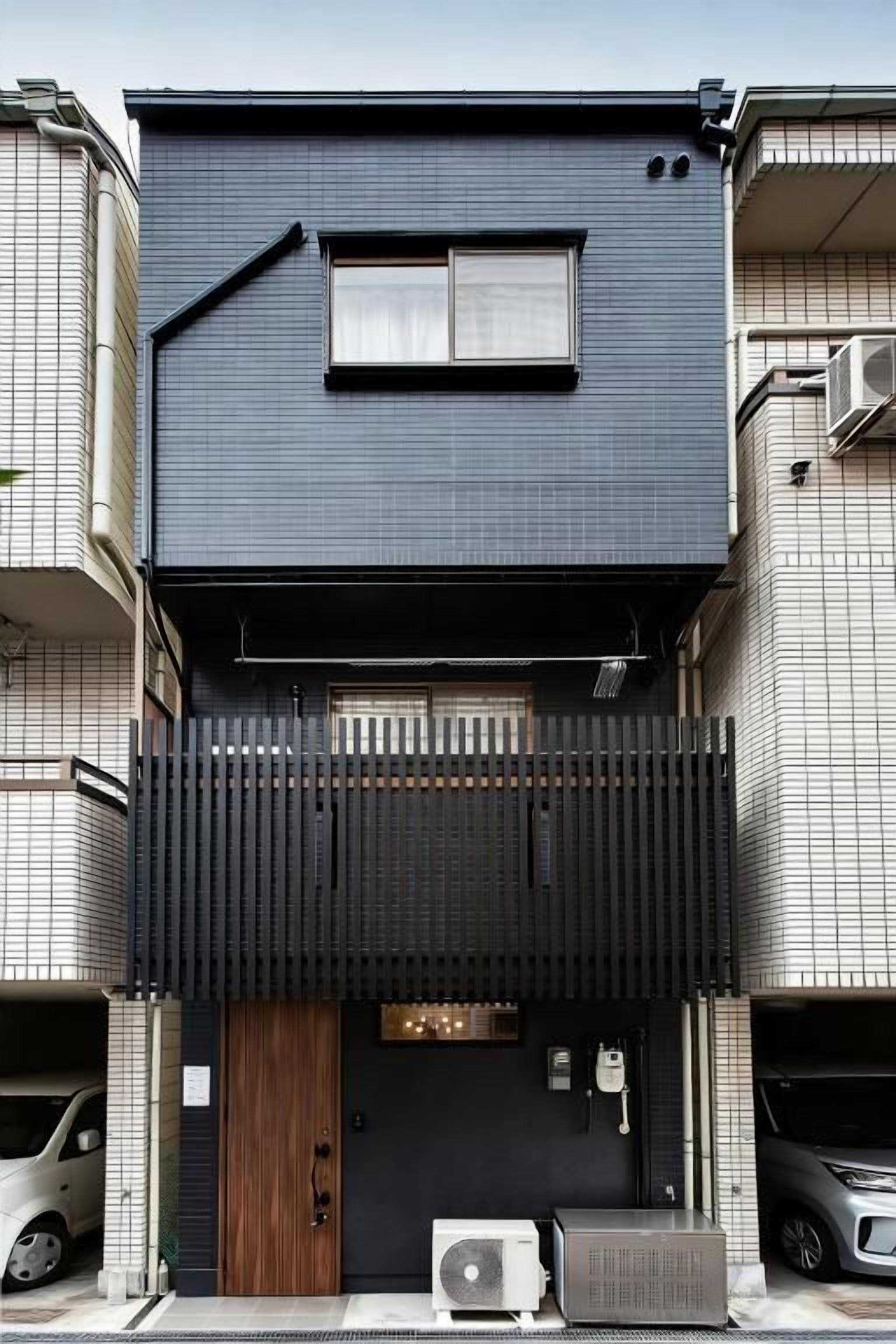 Un edificio moderno de dos pisos con fachada oscura, puerta de madera y una ventana pequeña.