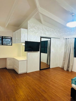 A room with a wooden floor, white walls, a flat-screen TV mounted on the wall, a desk with drawers, a wardrobe, and a sliding door.