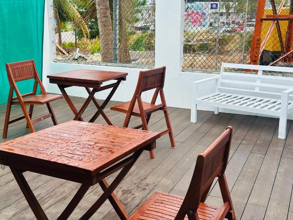 An outdoor seating area with wooden tables and chairs, a white bench, and a view through a chain-link fence.