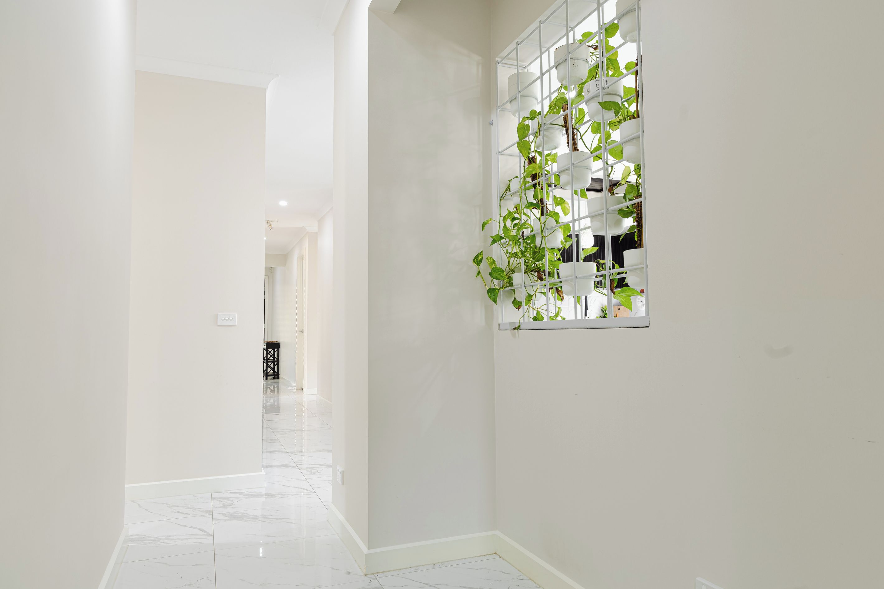 Un couloir avec un sol carrelé de carreaux blancs, un mur blanc et une fenêtre ornée d’une plante.