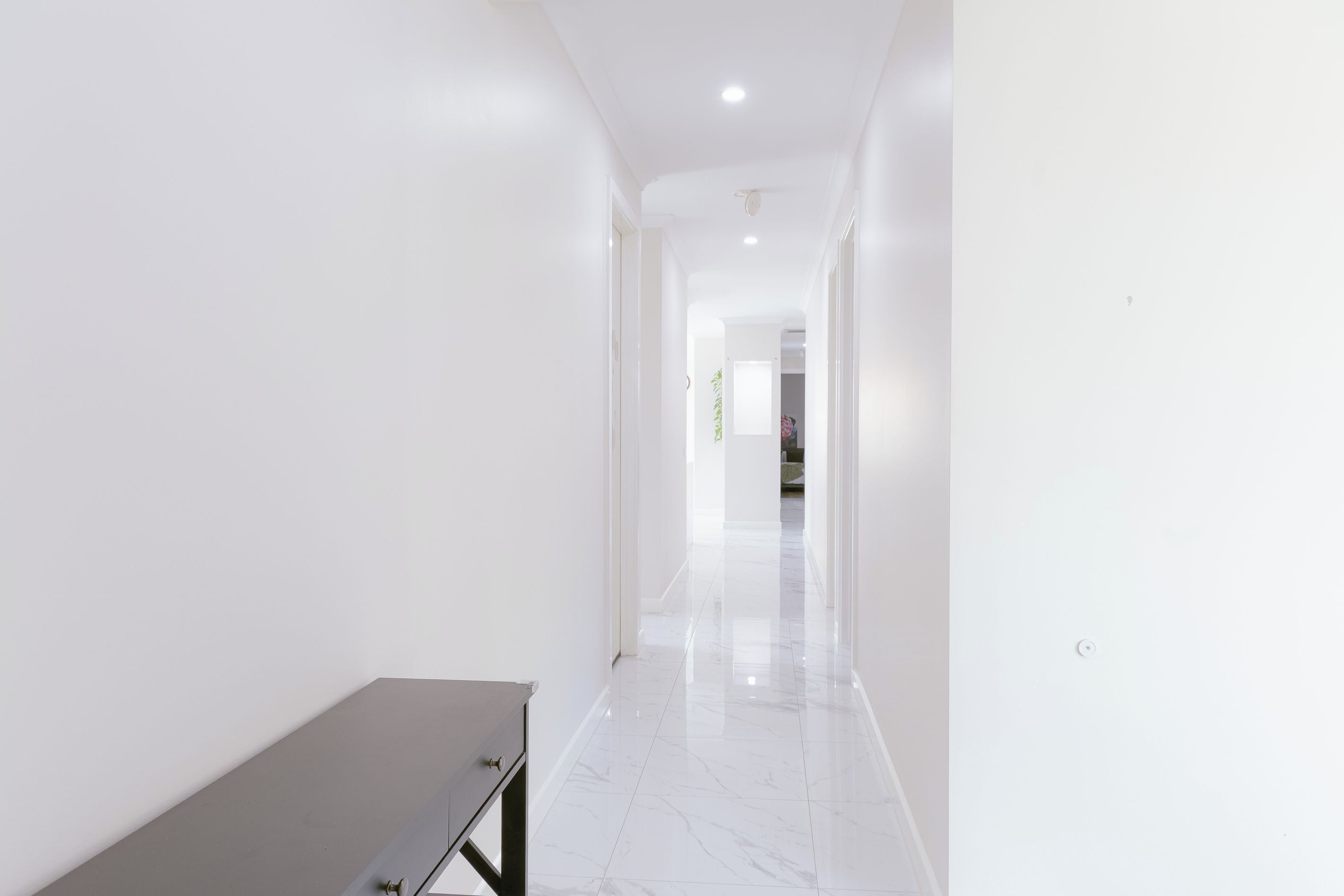 A hallway with white walls and a marble floor, leading to a room with a bed.