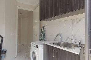 A compact kitchen with a built-in washing machine, a sink, and a door leading to another room. - Willows Arena House | Pet-friendly (Melbourne)