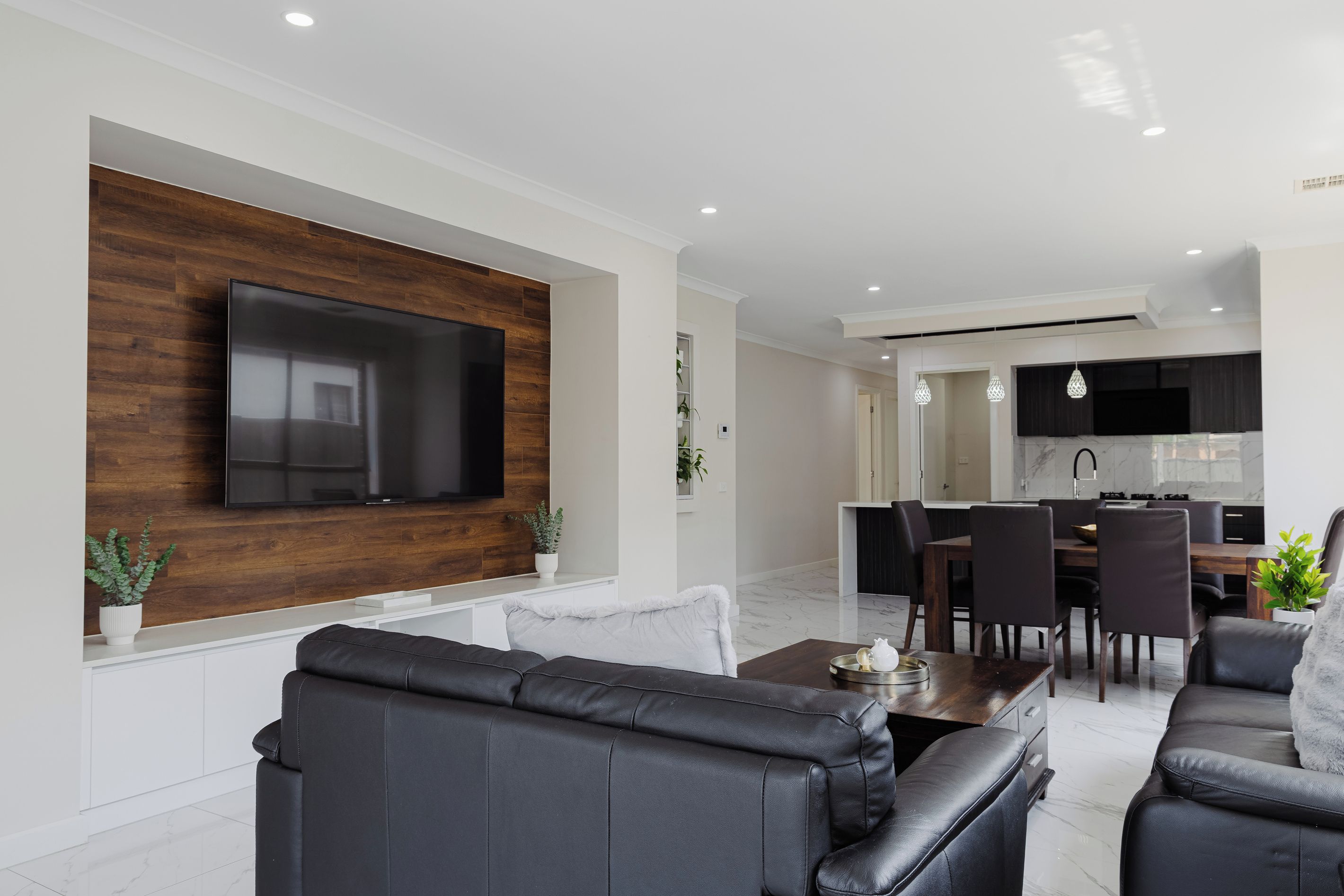 A modern living room with a flat-screen TV mounted on a wooden wall, a black leather sofa, and a dining area with a dark wooden table and chairs.