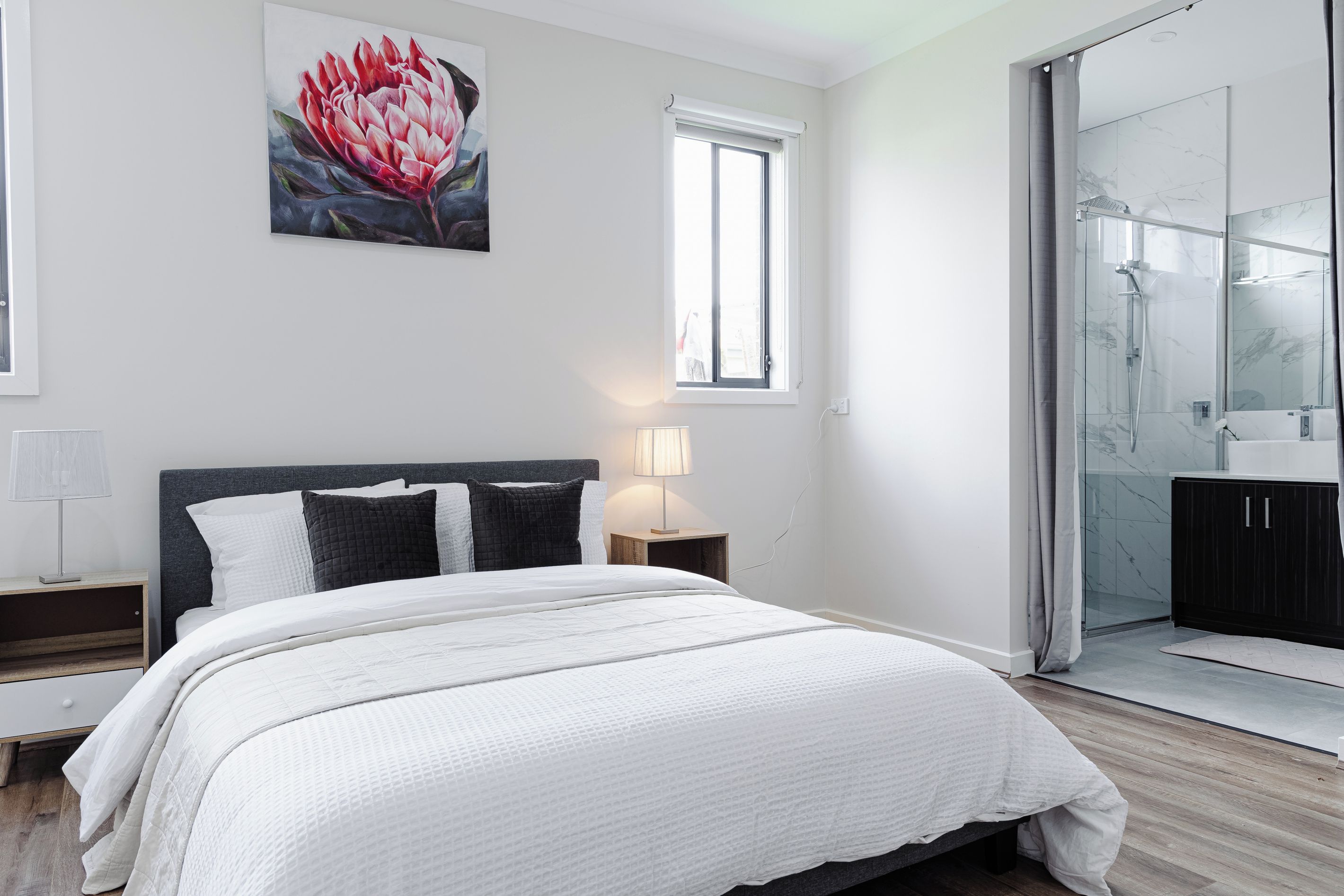 A modern bedroom with a large bed, a nightstand, a bedside lamp, a window, and a bathroom with a glass shower enclosure.