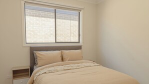 Un dormitorio con cama, mesita de noche, ventana con vista a un muro de ladrillo y una pared lisa.