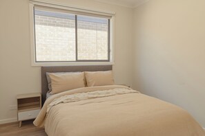 A bedroom with a bed, a nightstand, a window with a view of a brick wall, and a plain wall. - Willows Arena House | Pet-friendly (Melbourne)