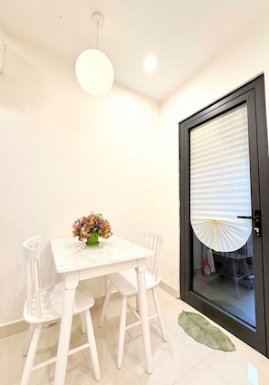 Uma sala de jantar com mesa e cadeiras brancas, luminária pendente e porta com persiana.