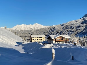 Paisaje nevado de montaña con dos edificios, uno con fachada de madera y otro con fachada color beige, ambos con techos cubiertos de nieve.