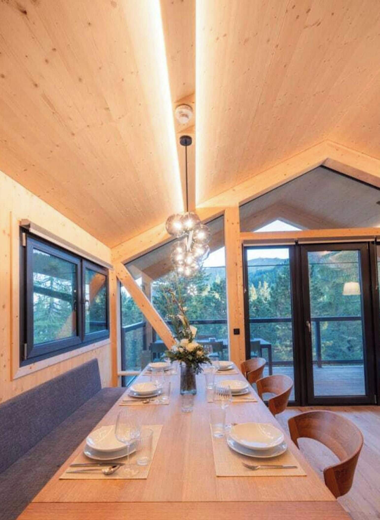 Un espace repas avec une longue table dressée pour quatre personnes, des murs en bois et de grandes fenêtres donnant sur l’extérieur.
