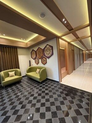 A hotel lobby with a checkered floor, two green sofas, a wall with decorative patterns, and a corridor leading to rooms.