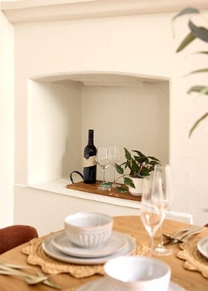A dining table with white plates, bowls, and a wine glass, set against a white wall with a shelf containing a wine bottle and glasses. - Sunny Bondi Junction 3BR | Backyard, BBQ & Bathtub (Bondi Junction)