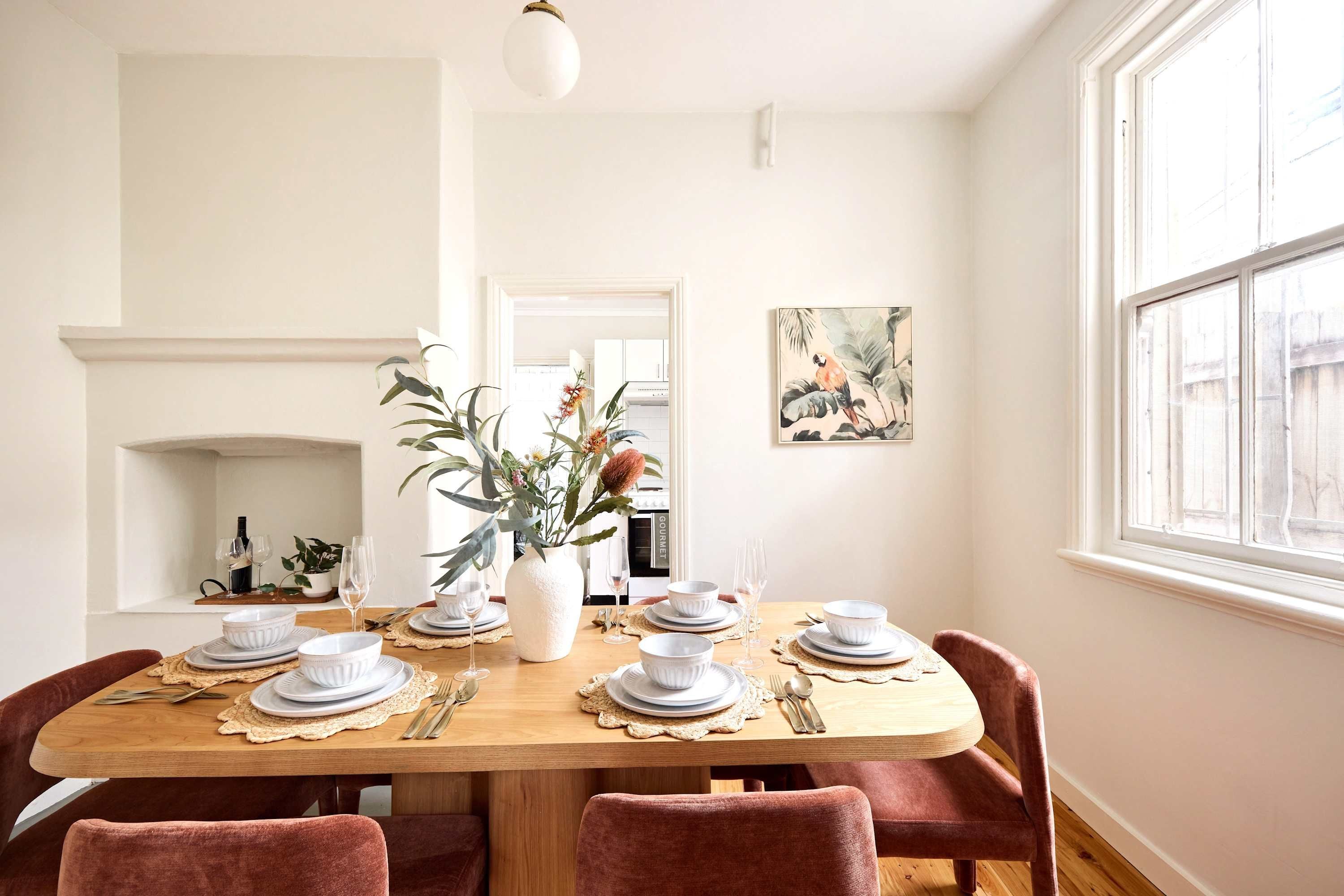 A dining room with a wooden table set for six, surrounded by red chairs, with a vase of flowers in the center.