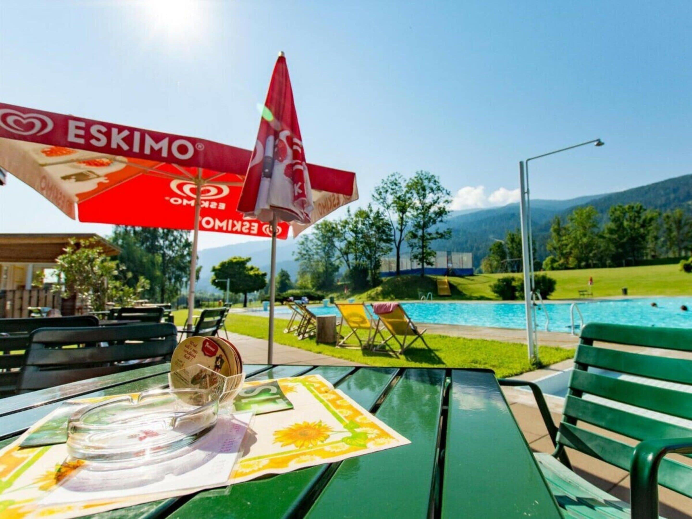 Een zwembadterras met een tafel, stoelen en een rode parasol met het logo 'ESKIMO'.