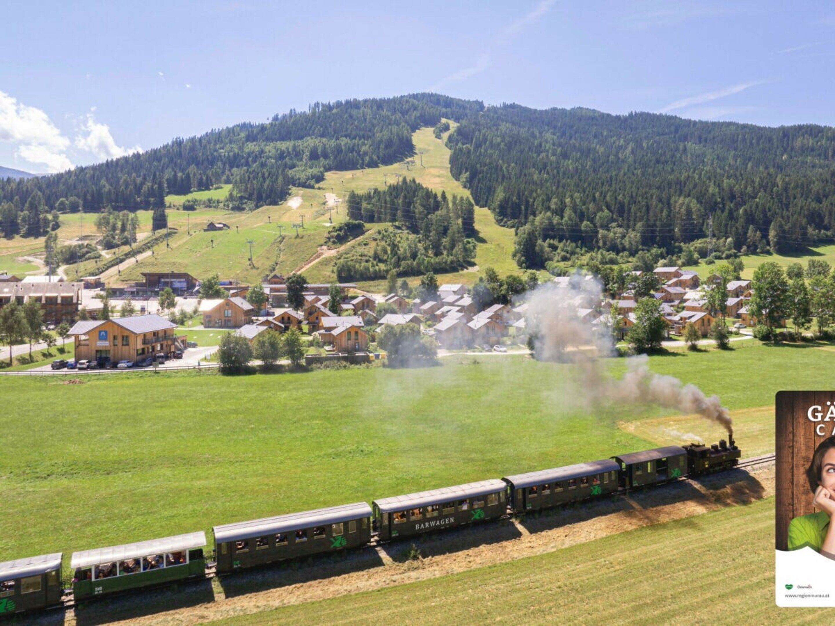 Ein Zug mit dem Schriftzug 'BARWAGEN', der durch eine ländliche Gegend fährt, im Hintergrund sind Häuser und ein bewaldeter Hügel zu sehen.