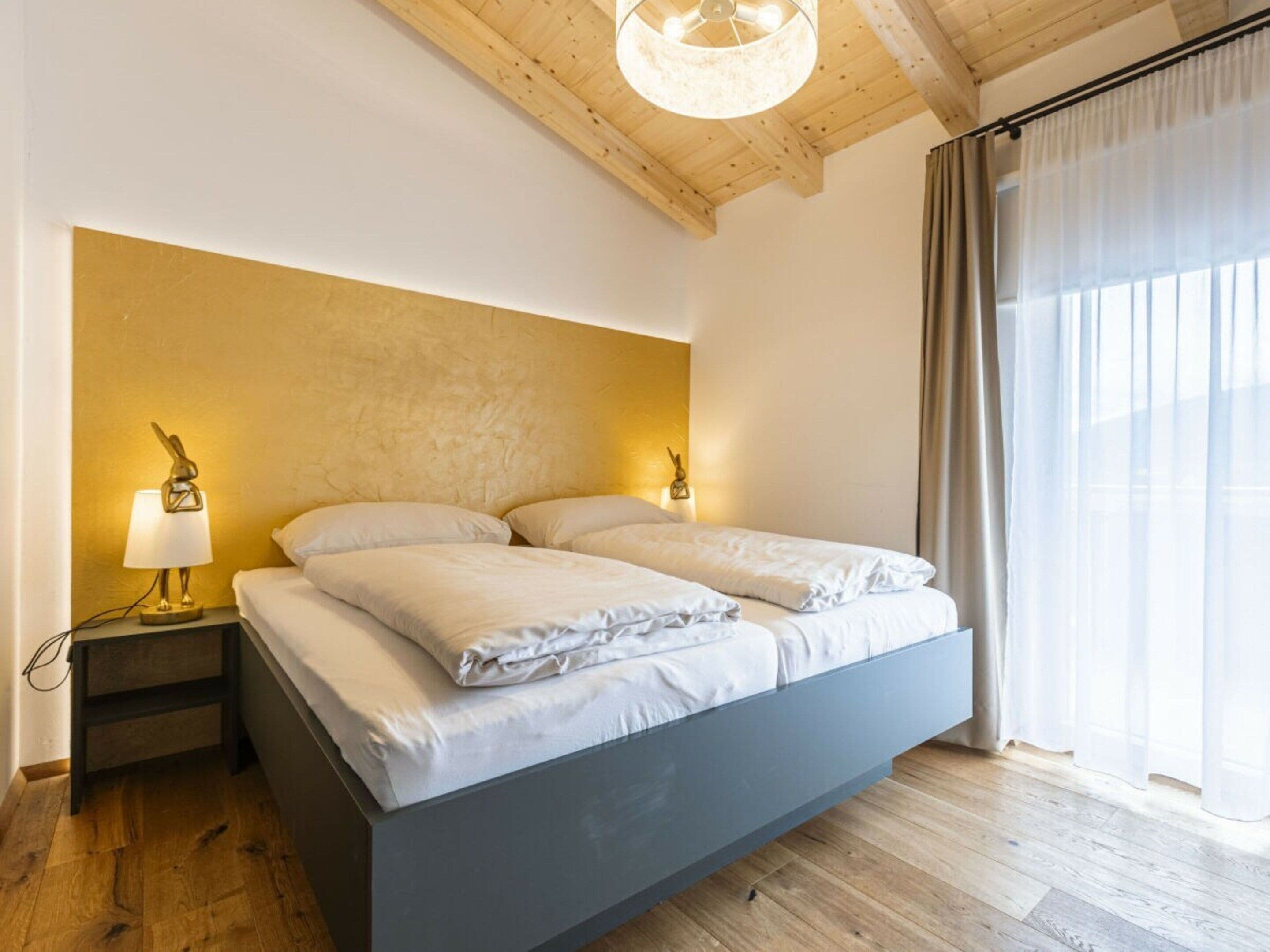 A bedroom with a bed, bedside lamps, a wooden ceiling, and a window with curtains.