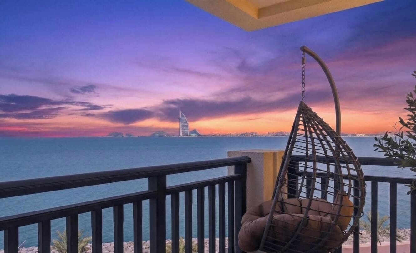 A balcony with a hanging chair overlooking a body of water and a distant building.
