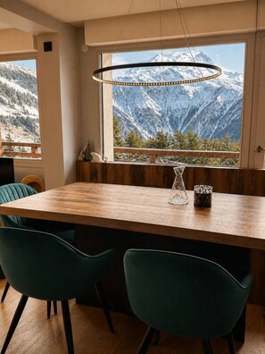A dining area with a wooden table, two teal chairs, a hanging light fixture, and a view of snow-capped mountains through a large window. - Dorion Lodge, Glacier View - Alto Society (Les Deux Alpes)