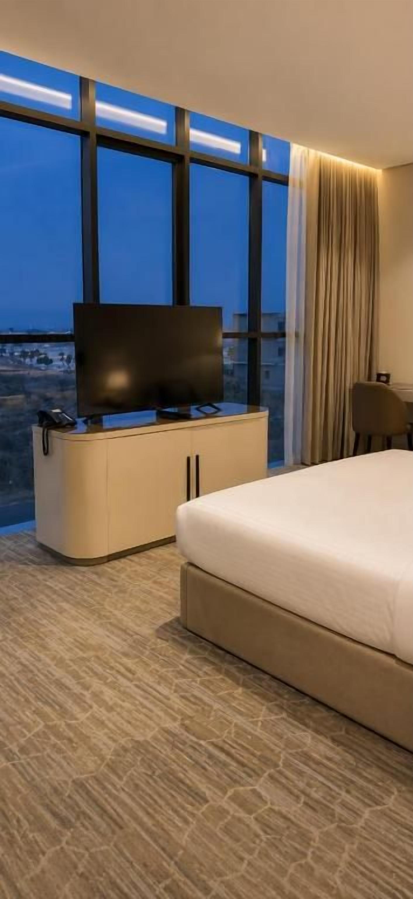 Habitación de hotel con cama, televisor en un mueble y vistas de la ciudad a través de amplios ventanales.