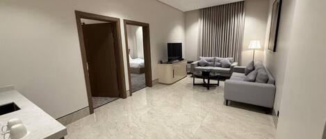 Una habitación de hotel moderna con piso de mármol, un sofá, un televisor, una mesita de noche y una lámpara.