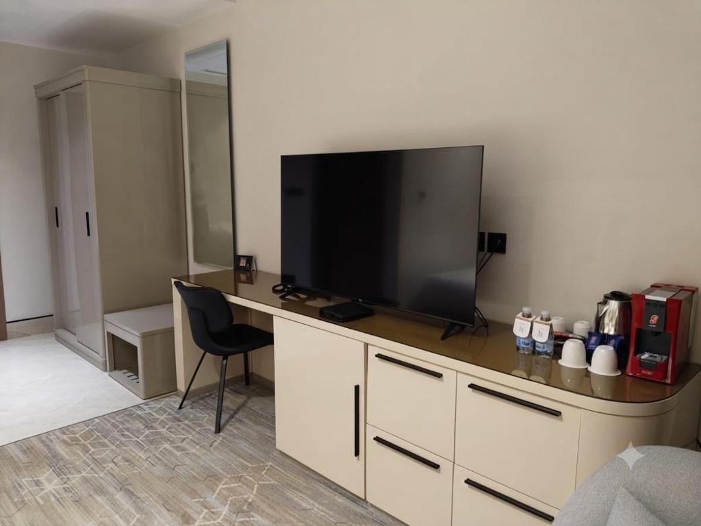 A hotel room with a flat-screen TV, a desk with a chair, a minibar, and a large mirror.