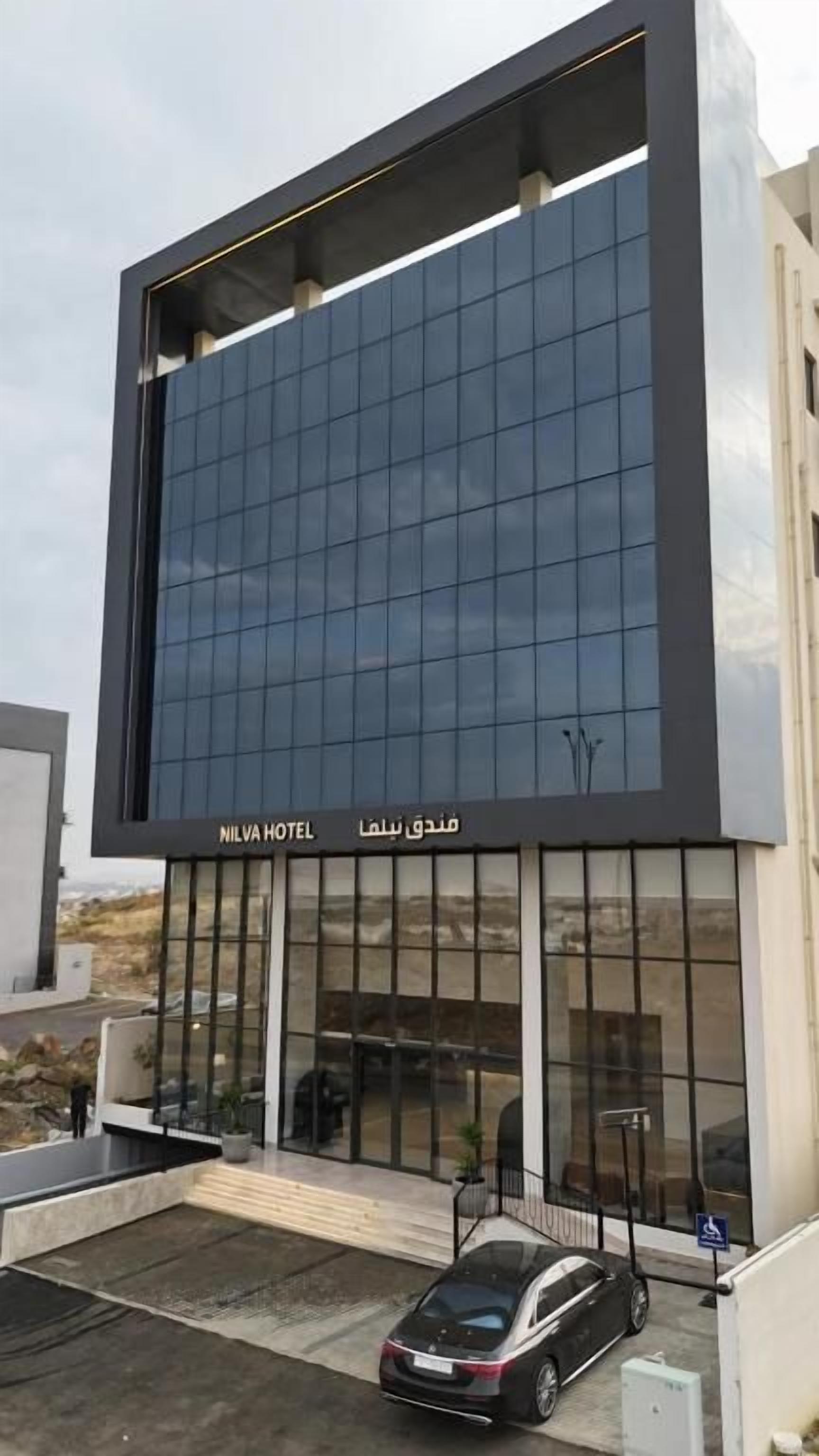 A modern hotel building with a large glass facade, a car parked in front, and a sign reading 'NILVA HOTEL'.