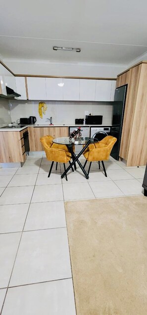 A modern kitchen with white cabinets, a black refrigerator, and a glass table with two yellow chairs. - Lovely One-Bedroom Unit with Pool and Gym Facilities in Stand (Cape Town)