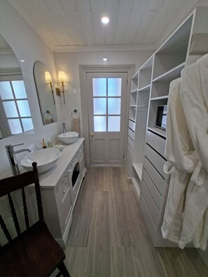 Un baño con un lavabo blanco, tocador y perchero. Se ve una silla de madera y una bata blanca colgada en un soporte.