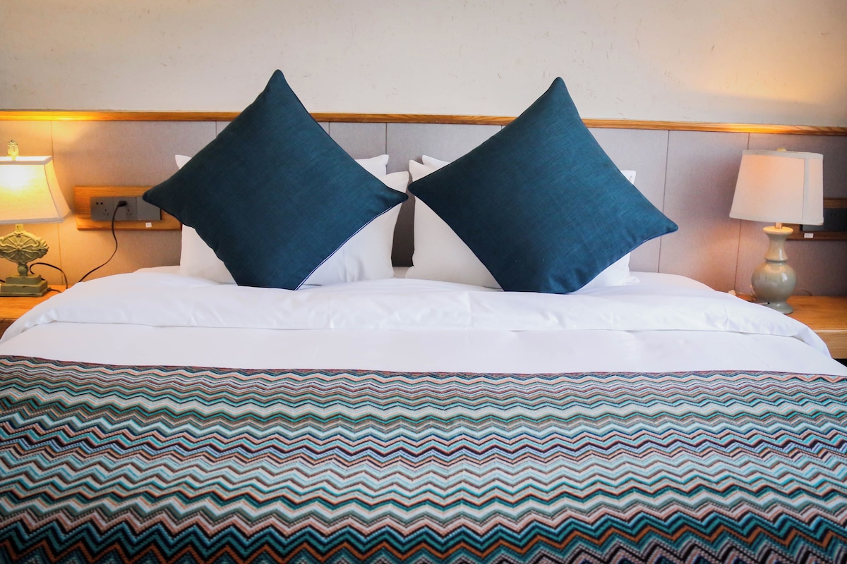 Habitación de hotel con cama, dos almohadas, colcha estampada, dos mesitas de noche con lámparas y un enchufe en la pared.