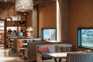 Interior de restaurante com móveis de madeira, luminárias de vime suspensas e vista pela janela.
