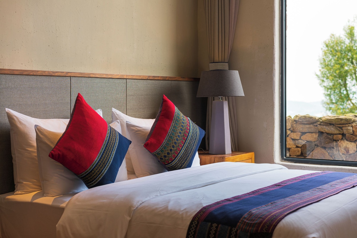 Una cama con ropa de cama blanca y almohadas decorativas, una mesita de noche con lámpara, y vistas a una pared de piedra y follaje a través de una ventana.