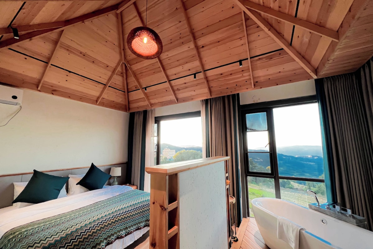 Un dormitorio con cama, mesita de noche, ventana grande y vistas al exterior.
