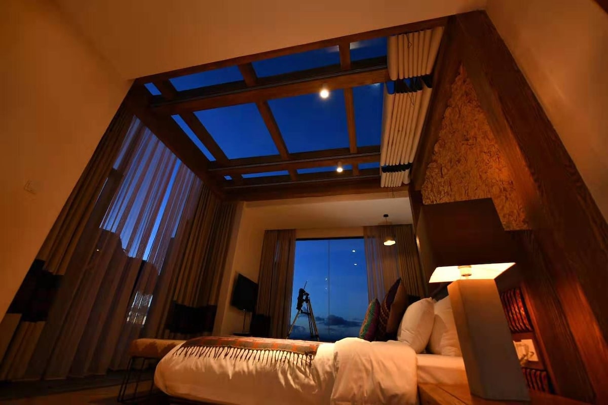 Habitación de hotel con una cama, una mesita de noche, una lámpara y vista al cielo a través de un tragaluz.