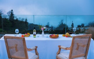 Mesa posta para dois com garrafa de vinho, copos e uma cesta de frutas.