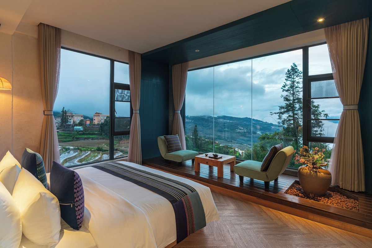 Habitación de hotel con una cama grande, dos sillones, una mesita y vistas a un paisaje montañoso.