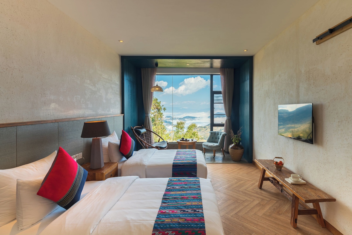 Habitación de hotel con dos camas, televisión, banco de madera y vista a las montañas.