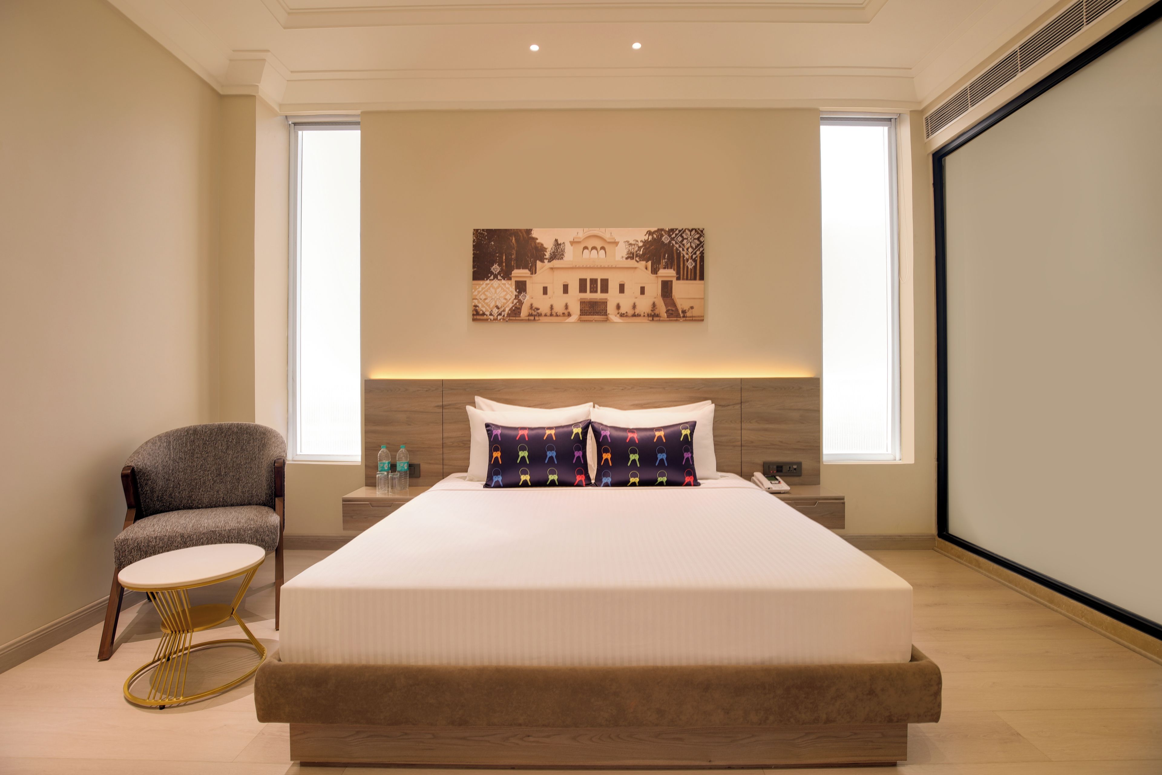 Habitación de hotel con una cama grande, una silla, una mesita pequeña y una pantalla proyectora montada en la pared.