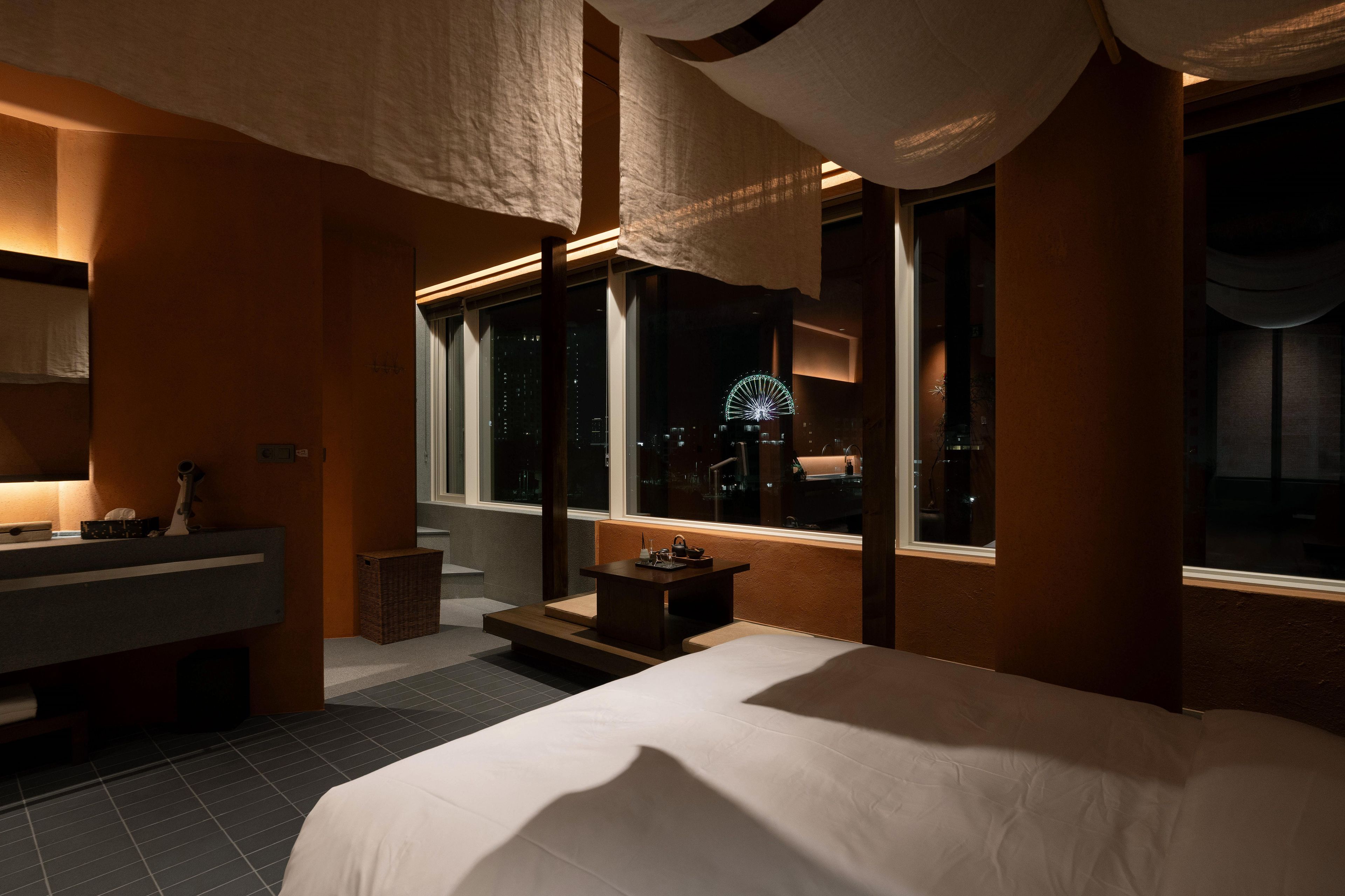 Habitación de hotel con una cama grande, baño con lavamanos y espejo, y ventanas amplias con vistas a la ciudad de noche.