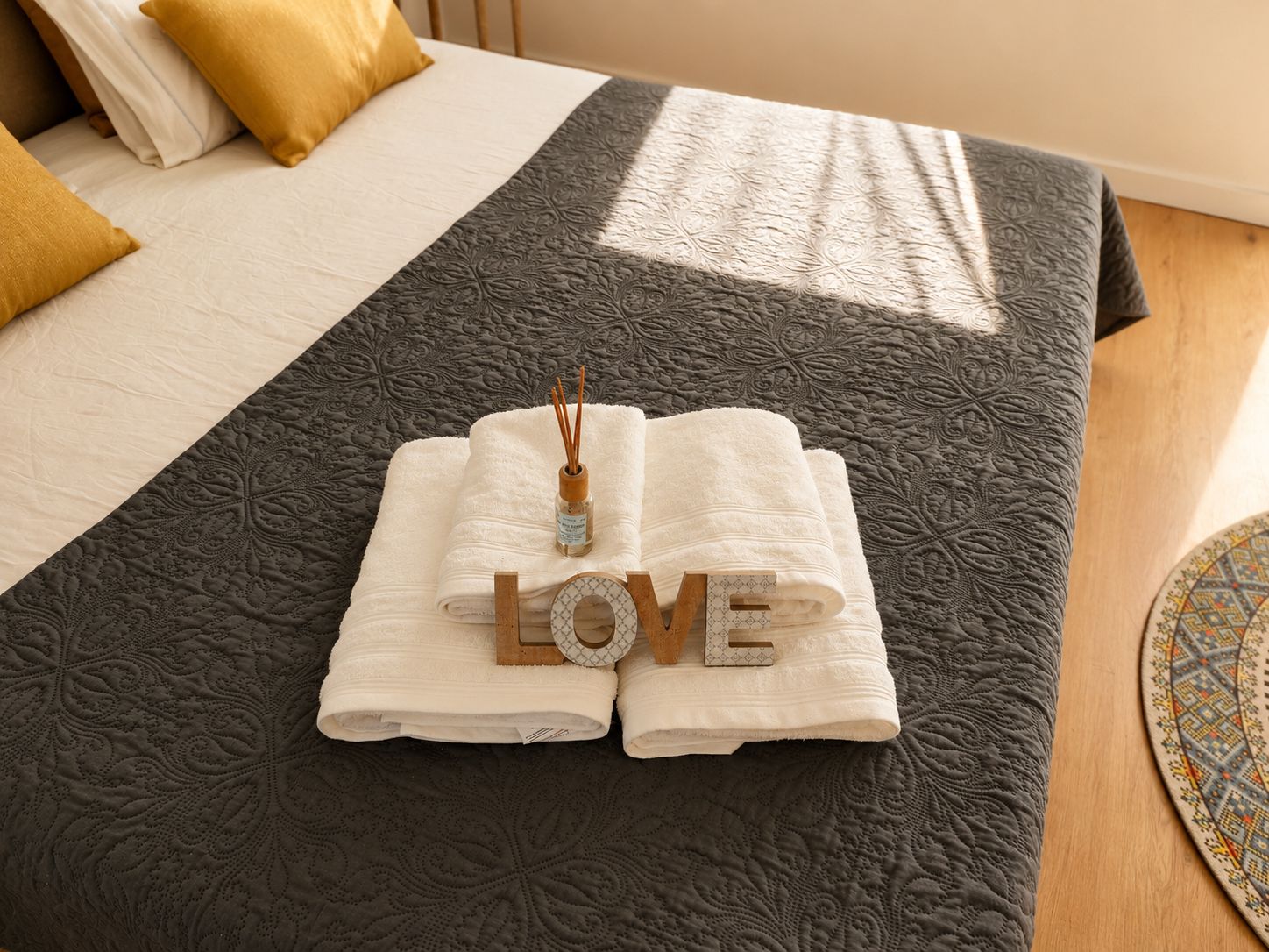 A neatly made bed with a dark patterned bedspread, two folded white towels, and a decorative item spelling 'LOVE' on the bed.