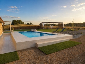 Una piscina rectangular amb una terrassa de pedra, dues gandules, una zona de seients coberta i un paisatge amb gespa al fons.