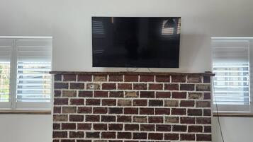 A room with a brick fireplace, a mounted flat-screen TV, and two windows with blinds.