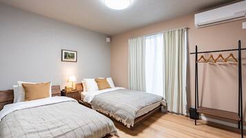 A hotel room with two beds, a bedside table with a lamp, a wall-mounted air conditioner, and a window with curtains.
