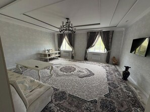 A room with a patterned carpet, a white sofa, a small table, a chandelier, and a window with curtains. - 2-bedroom apartment in adorable Bukhara with AC comfort (Bukhara)