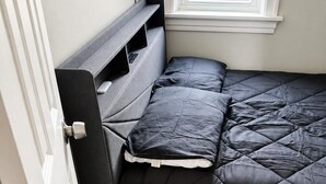 Una cama compacta con cabecera y piecera oscuros, colchón acolchado y ventana en lo alto.