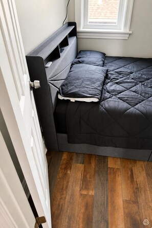 Een compact bed met een donker hoofdeinde en voeteinde, een gewatteerd matras en een raam erboven.