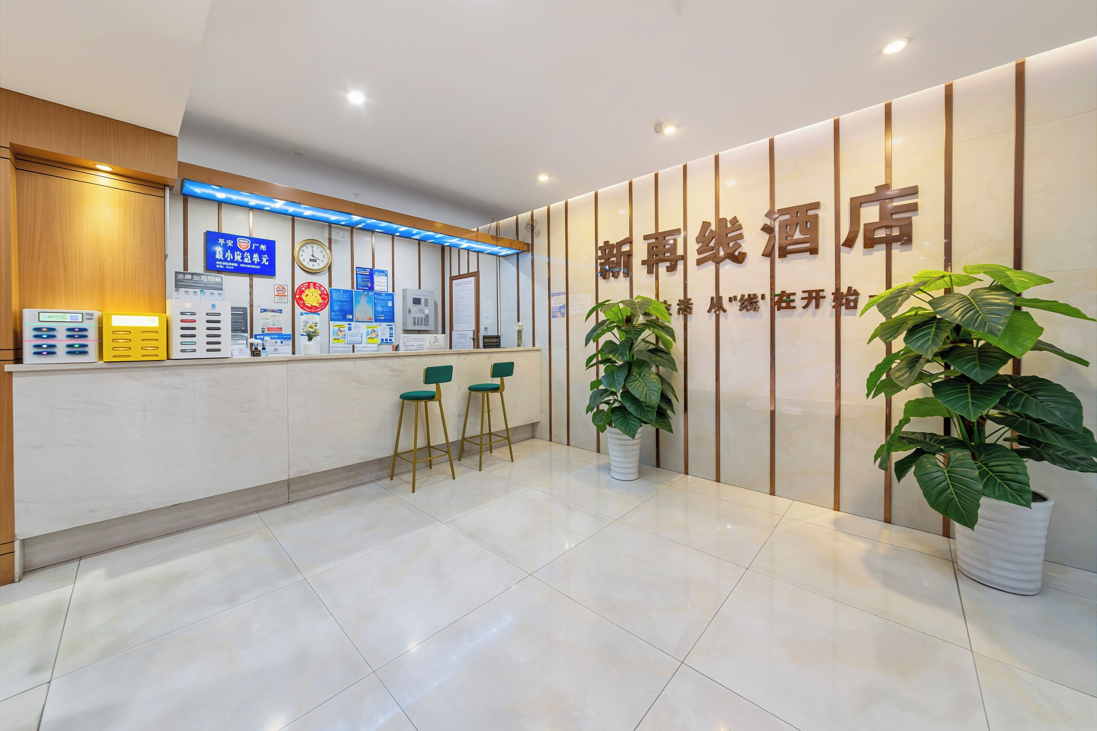 Área de recepção de hotel com balcão, cadeiras, relógio, cartazes informativos e plantas em vasos.